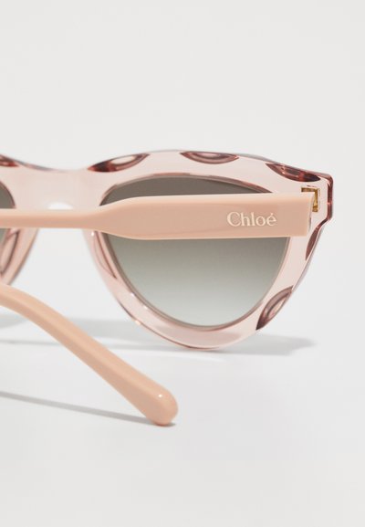 Lunettes de soleil roses, semi-transparentes, avec des verres ronds et une légère forme œil de chat. Accentuées par une monture rose clair et une marquage "Chloé" sur le côté.