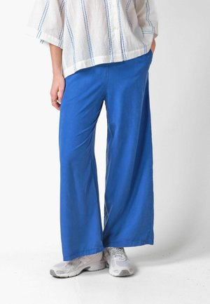 Persona che indossa pantaloni larghi di un brillante blu, scarpe da ginnastica bianche e una camicia bianca con sottili strisce blu, in piedi con le mani in tasca.