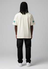 T-shirt en coton beige à coupe décontractée, manches courtes, et graphique turquoise sur la manche gauche. Associé à un jean noir et des baskets blanches.