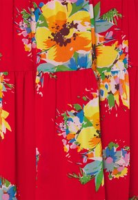 Roter Stoff mit einem bunten Blumenmuster, das große gelbe, orangefarbene und blaue Blumen zeigt. Das Design umfasst Falten und unterschiedliche Texturen.