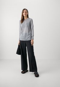 Grijze lange mouwen top met een subtiele glans, gecombineerd met zwarte wijde broek en stevig zwart sandaal, terwijl ze een gestructureerde zwarte handtas vasthoudt.