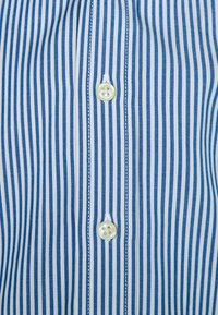 Niebieska i biała koszula w paski, z pionowymi paskami, z zapięciem na guziki i gładką bawełnianą teksturą. Dwa guziki widoczne na listwie.