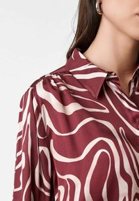 Woman wearing maroon buttoned blouse with white abstract swirl pattern and silver hoop earring, visible from neck to shoulder.