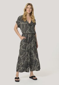 Svart och krämfärgad jumpsuit med bladmönster, V-ringning, korta ärmar, insvängd midja och vida byxor, stylad med svarta sandaler.