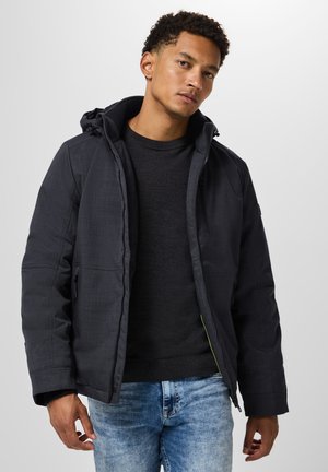 Zwarte gewatteerde jas met een capuchon, voorzien van een rits en zichtbare stiksels. Draagt over een donkere trui en blauwe jeans. Eenvoudig ontwerp.