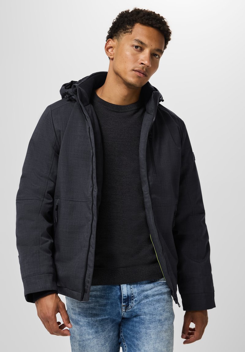 Black padded jacket with a hood, featuring a zip closure and visible stitching. Worn over a dark sweater and blue jeans. Simple design.