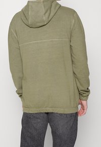 Sudadera color verde oliva con capucha ajustable, puños acanalados y dobladillo. Presenta una parte posterior sin costuras y una textura suave. Combinada con jeans grises.