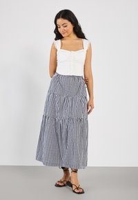 Hvit crop top med blonderemmer og knapper, kombinert med et svart-hvitt rutet, lagdelt maxiskjørt. Enkle svarte sandaler med stropper.
