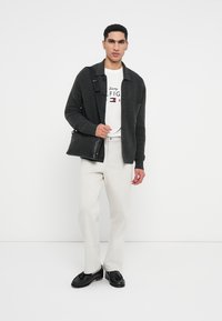 Fekete bőr táska, szürke kötött kardigán, fehér grafikás póló, krém színű nadrág, fekete cipő. A modell egy fehér háttér előtt áll.