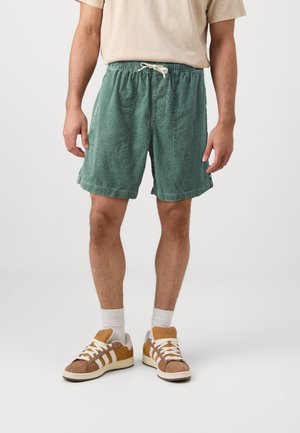 Homme portant un short vert texturé avec des poches devant, un t-shirt beige, des chaussettes blanches montantes et des baskets beige et blanches, debout sur fond blanc.