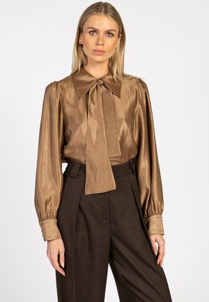 Bronzen metalen blouse met een grote kraag en nektie, gecombineerd met hooggetailleerde gestructureerde bruine broek. Lange mouwen met aansluitende manchetten.
