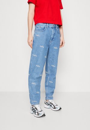 Osoba w jasnoniebieskich dżinsach z białymi haftowanymi logo, czerwonym topie z krótkim rękawem oraz białych i czarnych butach sportowych stojąca na jednolitym tle.