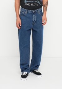 Baggy blå denimjeans med lys vask, store lommer og knappelukking, kombinert med svarte lerretsko og en svart grafisk T-skjorte.