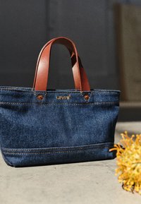 Τσάντα ώμου από τζιν με καφέ δερμάτινες λαβές, χρυσό λογότυπο και αντίθετες ραφές. Συμπαγής σχεδίαση με ενισχυμένες άκρες και ελάχιστο υλικό.