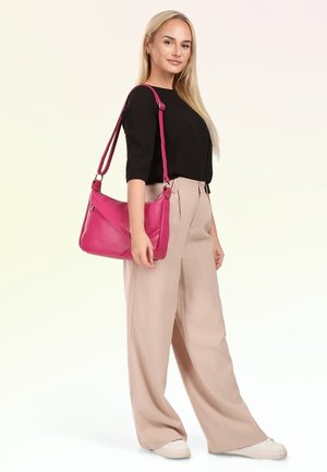 Femme blonde portant un haut noir et un pantalon beige à jambes larges, portant un sac à bandoulière rose vif, debout devant un fond clair.