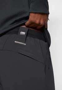 Melni sporta šorti ar elastīgu jostasvīru, uz kuriem ir uzlīme "ciele ATHLETICS" un neliela atstarojoša detaļa tuvu kabatai.