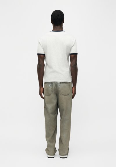 Άντρας που κοιτάζει προς τα πίσω, φοράει λευκό μπλουζάκι με μαύρο περίγραμμα και άνετο ξεθωριασμένο πράσινο τζιν παντελόνι, στέκεται σε απλό λευκό φόντο.