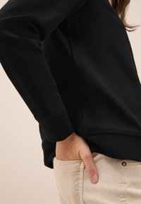 Felpa nera con tessuto texture e una vestibilità leggermente ampia, abbinata a pantaloni beige chiaro. La mano appoggiata in tasca ne mostra i dettagli.