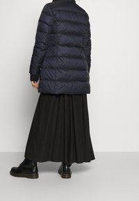 Marineblauwe pufferjas met doorgestikte patroon, lange mouwen, hoge kraag en een ontspannen pasvorm, gecombineerd met een vloeiende zwarte rok en zwart veterschoenen.