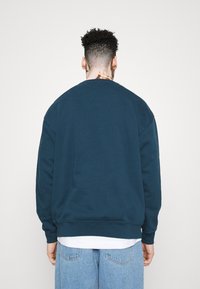 Tmavě modrý svetr s kulatým výstřihem, volným střihem a ribovanými manžetami. Materiál vypadá měkce, bez viditelných vzorů nebo log.