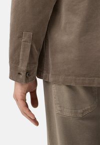 Brun kordflorskjorta med knäppte ärmar, som visar ribbad textur. Hand något utsträckt, med slät hud mot det ljusa tyget på byxorna.