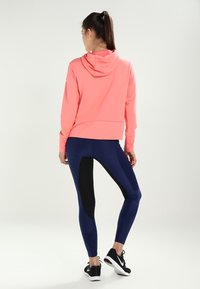 Roze hoodie met een capuchon en een buidelzak, gecombineerd met marineblauwe leggings met zwarte zijkanten. Het materiaal lijkt rekbaar en sportief.
