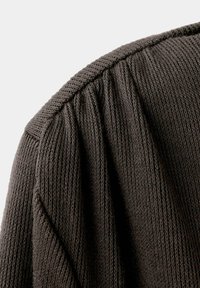 Gros plan d’un tissu tricoté brun foncé montrant une texture côtelée détaillée et des coutures froncées le long d’une couture d’épaule.
