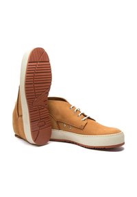 Chaussures en cuir marron avec des lacets blancs et une semelle en caoutchouc crème. Tige texturée avec œillets en métal et semelle extérieure à motif rouge.