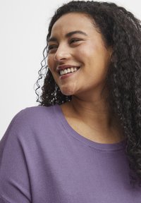Maglione viola lavorato a maglia con una vestibilità comoda, scollatura rotonda e dettagli a coste. La modella ha i capelli ricci e sorride.