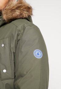Mężczyzna w oliwkowej zimowej kurtce z futrzanym kapturem i okrągłą naszywką Jack & Jones na górnej części rękawa.