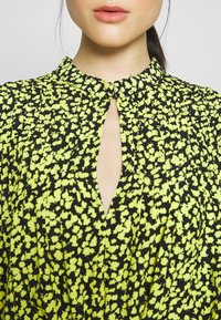 Černé šaty s živým žlutým květinovým vzorem, s vysokým límcem a detaily na krku ve tvaru klíče, z lehkého materiálu.