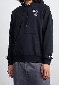 Svart huva sweatshirt i mjukt tyg med en känguruficka, silverfärgade dragsnören och en vit grafisk logo på vänster bröst.