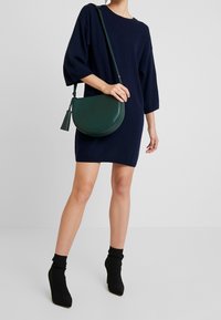 Ciemnozielona skórzana torebka na krzyż o półokrągłym kształcie, z detalem w postaci frędzli i regulowanym paskiem, zestawiona z granatową, oversize'ową sukienką oraz czarnymi botkami.