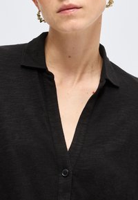 Κοντινό πλάνο προσώπου που φοράει μαύρο πουκάμισο με γιακά και βαθύ V-λαιμόκοψη, καθώς και χρυσούς στριφογυριστούς κρίκους σκουλαρίκια.