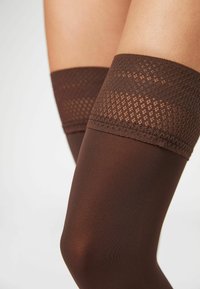 Bas collants marron montants jusqu'à la cuisse avec une bande en dentelle texturée en haut et une finition lisse et opaque. Le matériau semble extensible et ajusté.