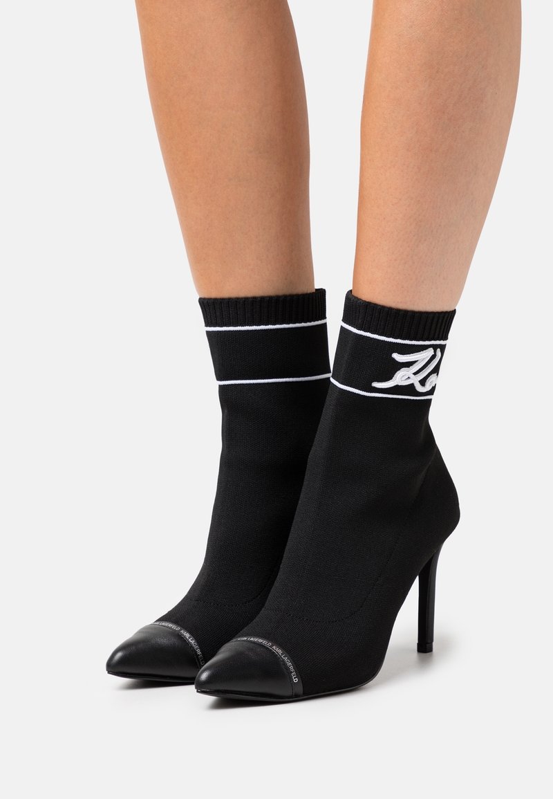 Botas de tobillo de punto negro con un diseño elegante, puntera afilada y tacón stiletto alto. Presentan puños acanalados y costuras de acento en blanco.