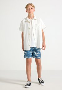 Camicia bianca a maniche corte con bottoni, pantaloni da bagno azzurri con motivo, scarpe da ginnastica nere, in piedi contro uno sfondo chiaro e uniforme.