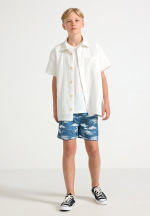 Witte overhemd met korte mouwen, blauwe gebloemde zwemshorts, zwarte sneakers, staande tegen een effen lichte achtergrond.