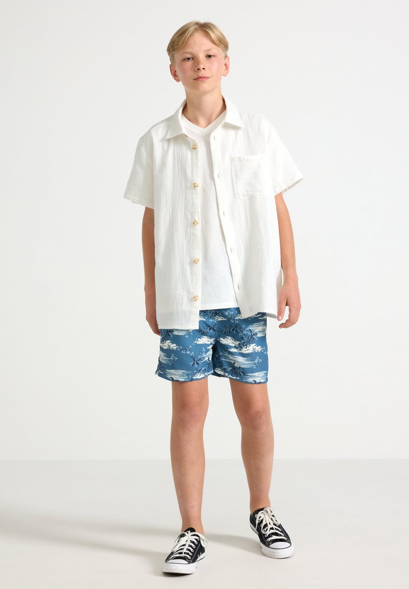 Camicia bianca a maniche corte con bottoni, pantaloni da bagno azzurri con motivo, scarpe da ginnastica nere, in piedi contro uno sfondo chiaro e uniforme.
