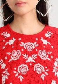 Top in pizzo rosso con ricami floreali bianchi, scollatura rotonda e un motivo testurizzato. Grandi orecchini geometrici completano il design.
