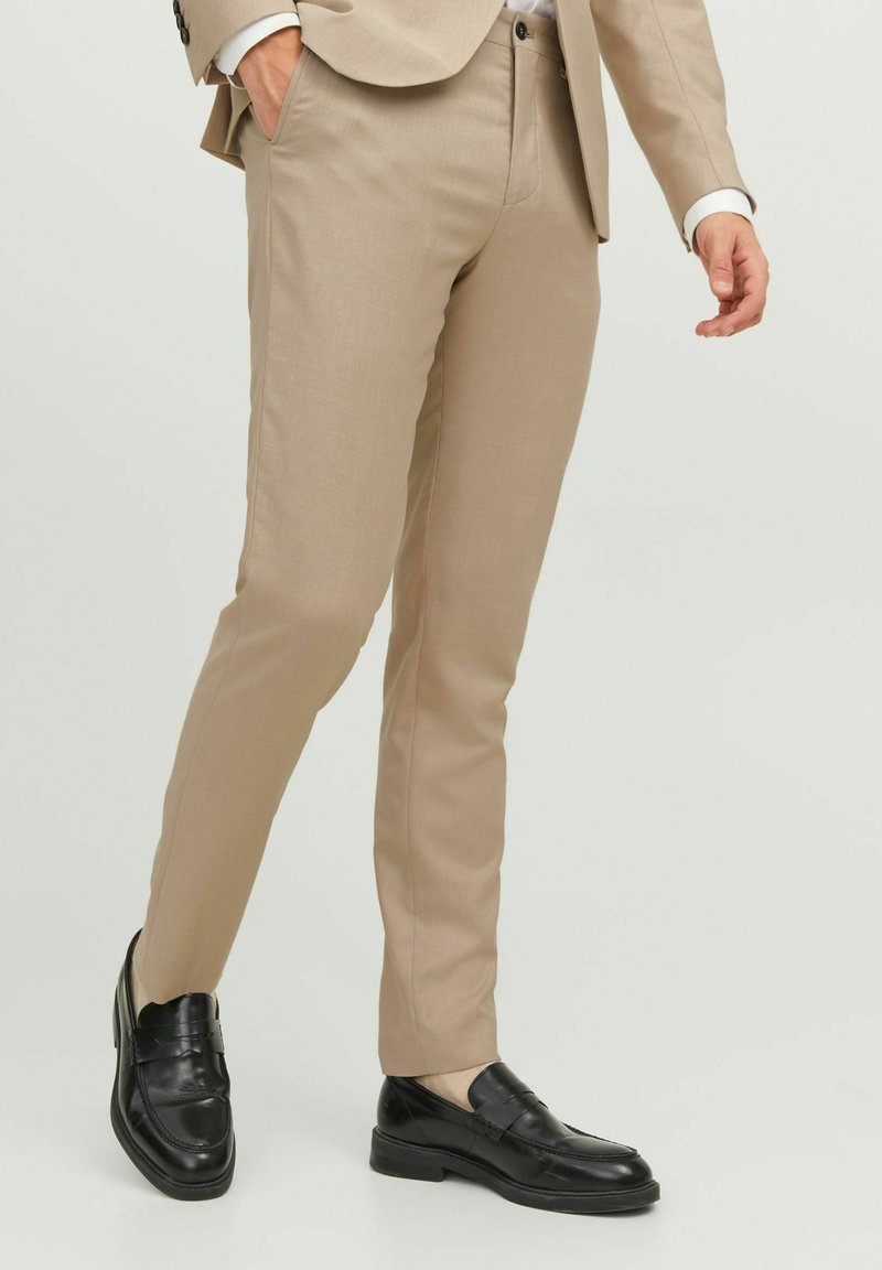 Beige maßgeschneiderte Hose aus glattem Stoff mit schmaler Passform, ausgestattet mit Seitentaschen und Biesen, kombiniert mit schwarzen Slippers.