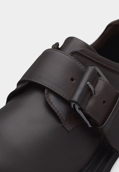 Σκούρο καφέ δερμάτινο παπούτσι με φαρδιά λουρίδα και μεταλλική αγκράφα σε απλό ανοιχτό φόντο.