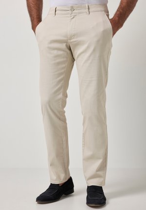 Homme portant un pantalon beige coupe droite avec poches latérales et des mocassins noirs, debout devant un fond clair uni.