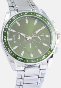 Relógio de aço inoxidável prateado com um mostrador texturizado verde, marcadores de horas brancos, três submostradores e um bezel verde. Bracelete de elos de metal.
