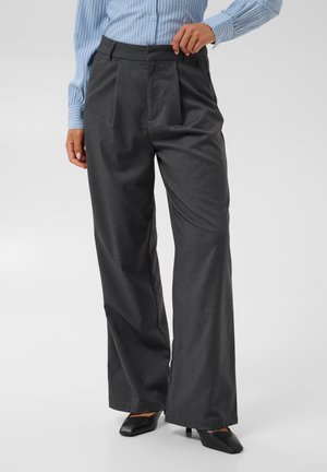 Pessoa vestindo calças largas cinza escuro, camisa azul clara às riscas e saltos pretos pontiagudos, em pé contra um fundo simples.