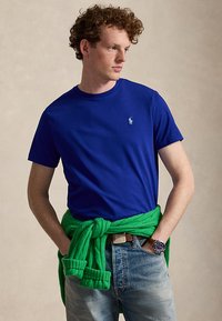 Jeune homme aux cheveux bouclés portant un t-shirt bleu, un pull vert noué autour de la taille, un jean clair et une montre-bracelet, regardant sur le côté.