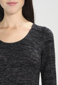 Donna che indossa un top grigio scuro a maniche lunghe con scollo rotondo, che mostra il collo e la parte superiore del petto.