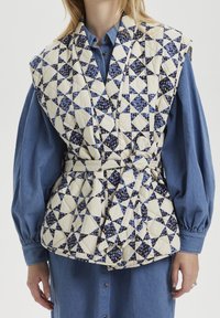 Vadderad väst med ett geometriskt mönster i kräm, blått och marinblått. Har en bältad midja, breda slag och en lagerdesign över en jeansskjorta.