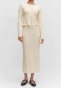 Conjunto de punto de dos piezas en beige claro, que incluye un cárdigan corto de manga larga con botones y una falda larga y ajustada. Unos zapatos planos completan el conjunto.