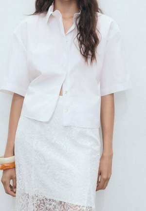 Femme portant une chemise blanche à manches courtes boutonnée et une jupe blanche en dentelle, avec des cheveux bouclés foncés et des bracelets à son poignet gauche.